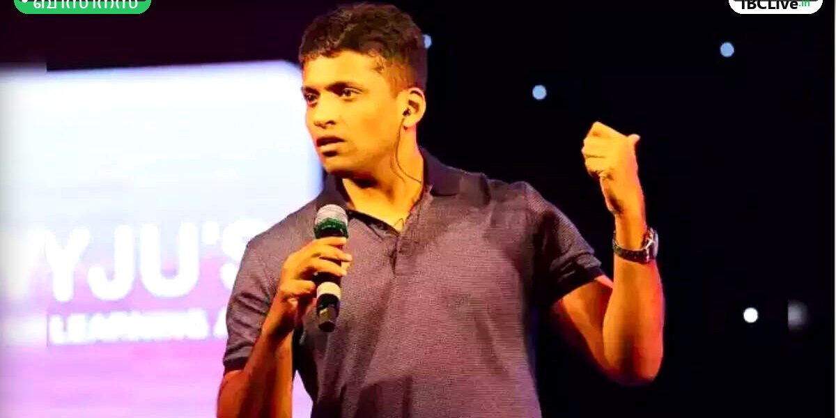 ബൈജു രവീന്ദ്രൻ മൈക്കിലൂടെ സംസാരിക്കുന്നു, ഒരു വലിയ സ്ക്രീനിന്റെ മുന്നിൽ