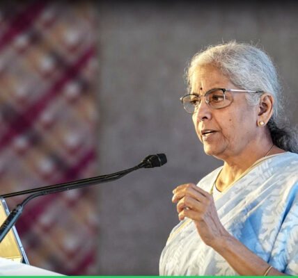കേന്ദ്ര ധനകാര്യ മന്ത്രി നിർമ്മല സീതാരാമൻ മാധ്യമങ്ങളോട് സംസാരിക്കുന്നു. പിന്നിൽ ഇന്ത്യൻ പതാകയുടെ ചിത്രീകരണം.
