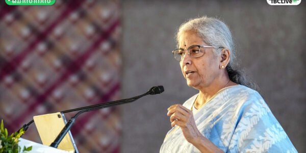 കേന്ദ്ര ധനകാര്യ മന്ത്രി നിർമ്മല സീതാരാമൻ മാധ്യമങ്ങളോട് സംസാരിക്കുന്നു. പിന്നിൽ ഇന്ത്യൻ പതാകയുടെ ചിത്രീകരണം.